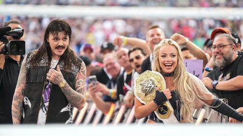 Dominik Mysterio and Liv Morgan look towards the wrestling ring. Liv Morgan holds the Women's World Championship on her shoulder.