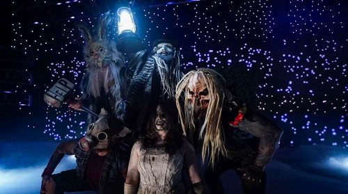 A group of wrestlers in various costumes and masks huddle together in the middle of the wrestling ring. The center figure, Uncle Howdy, holds a lit lantern aloft.